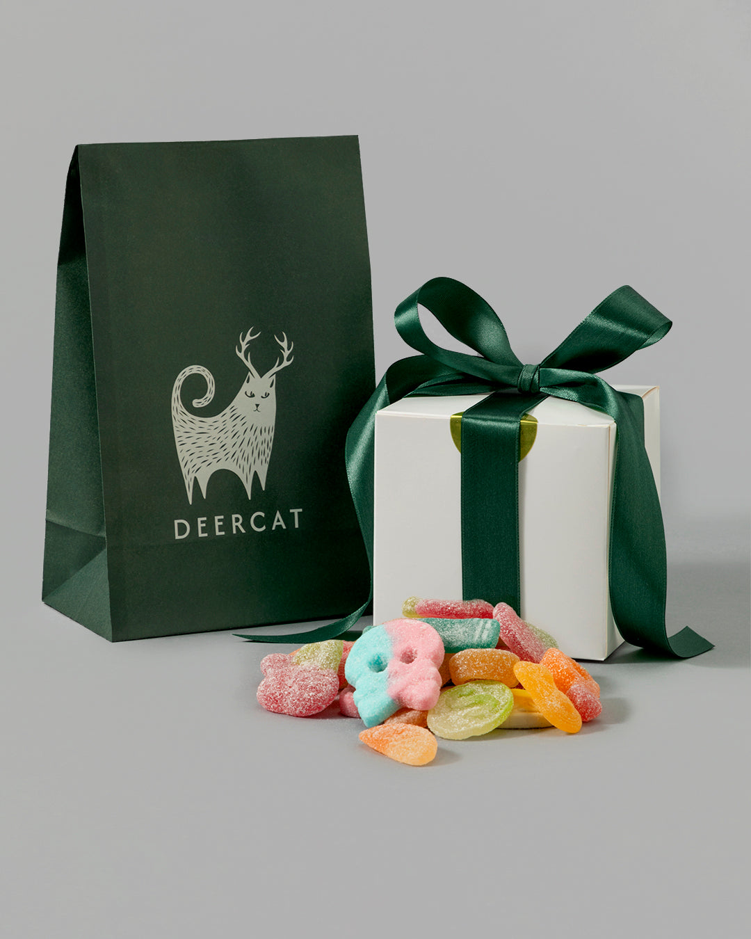Dark green gift bag with Deercat logo next to a white box with green ribbon and colorful candy pieces scattered nearby