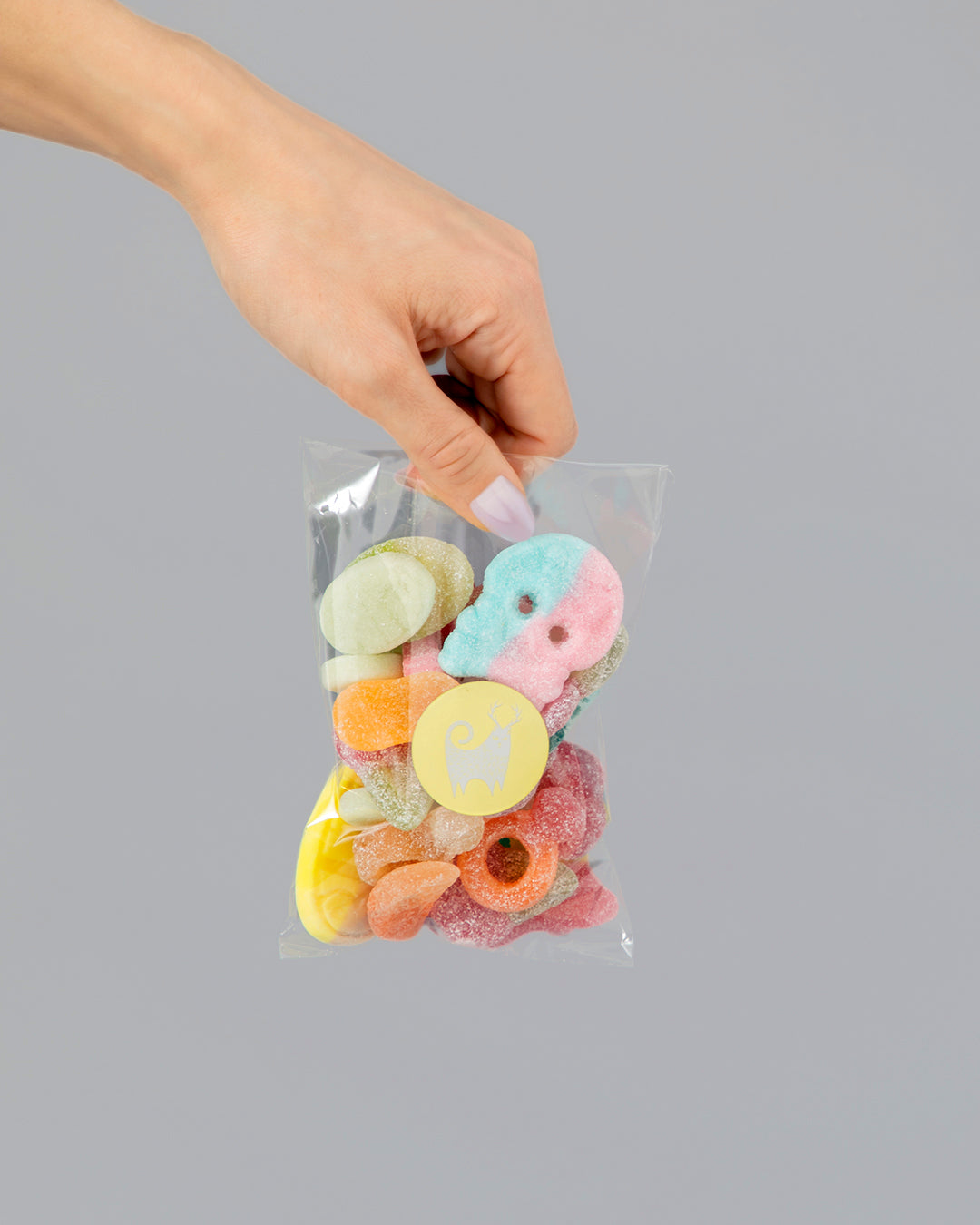 Hand holding clear bag filled with colorful vegan candy mix against a plain gray background