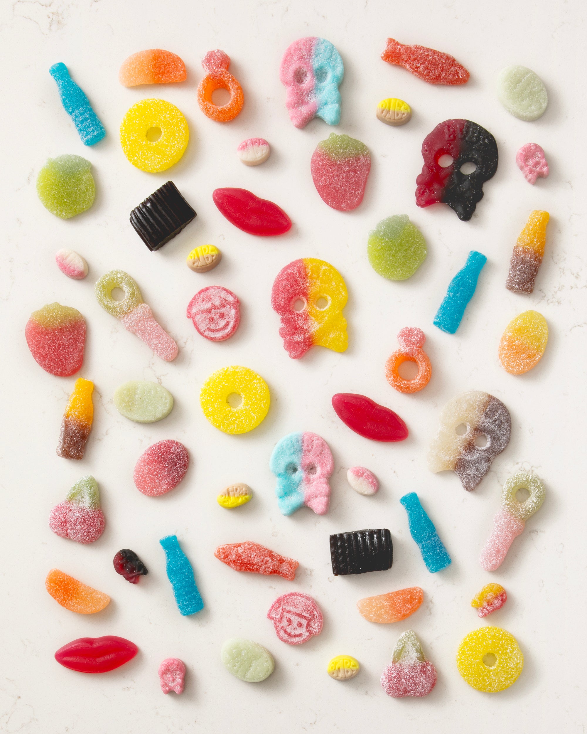 Colorful vegan candy mix with playful shapes like skulls, lips, bottles, rings, and fruit on a light surface.
