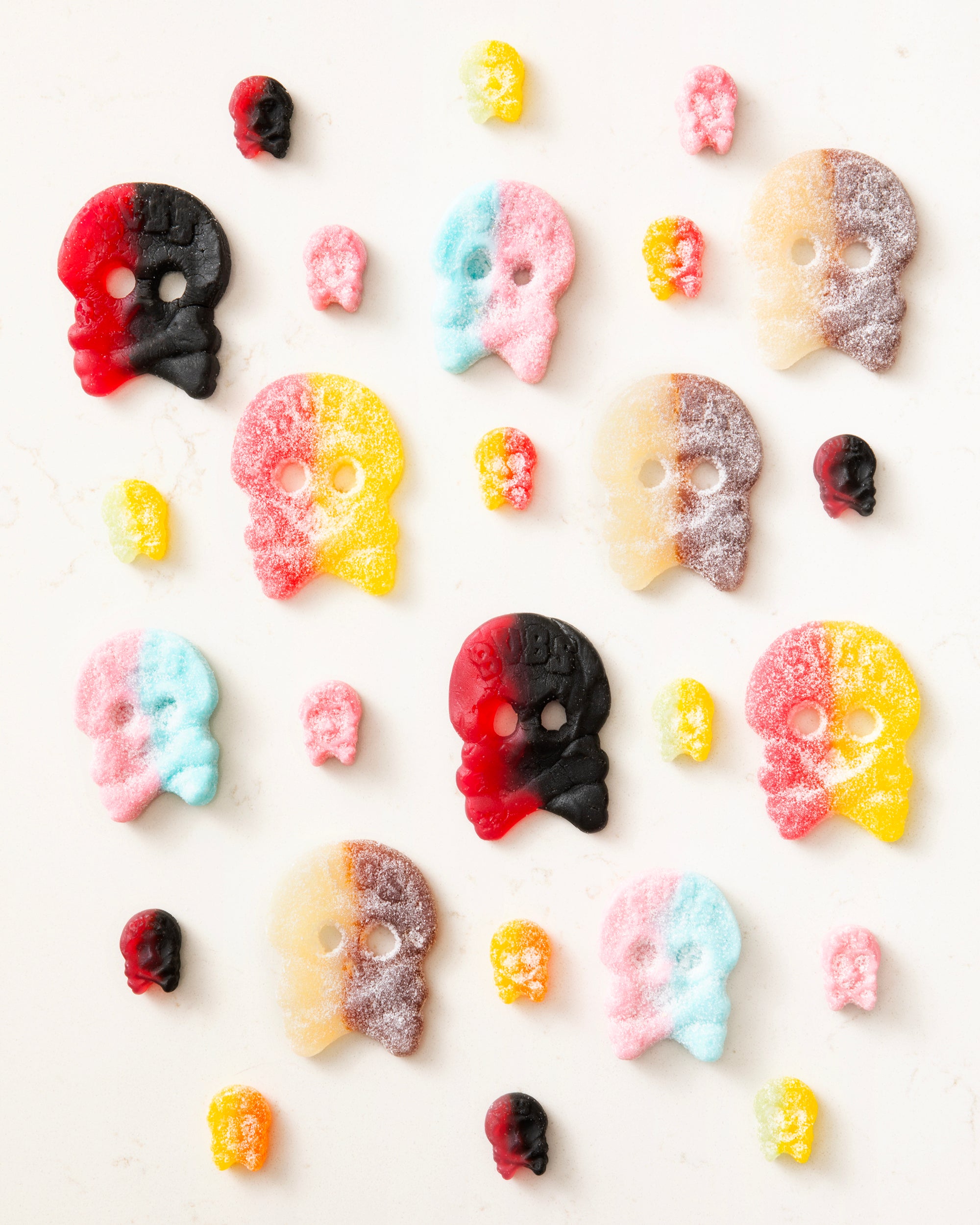 Colorful two-tone skull-shaped and gummy candies arranged on a white surface in a playful pattern