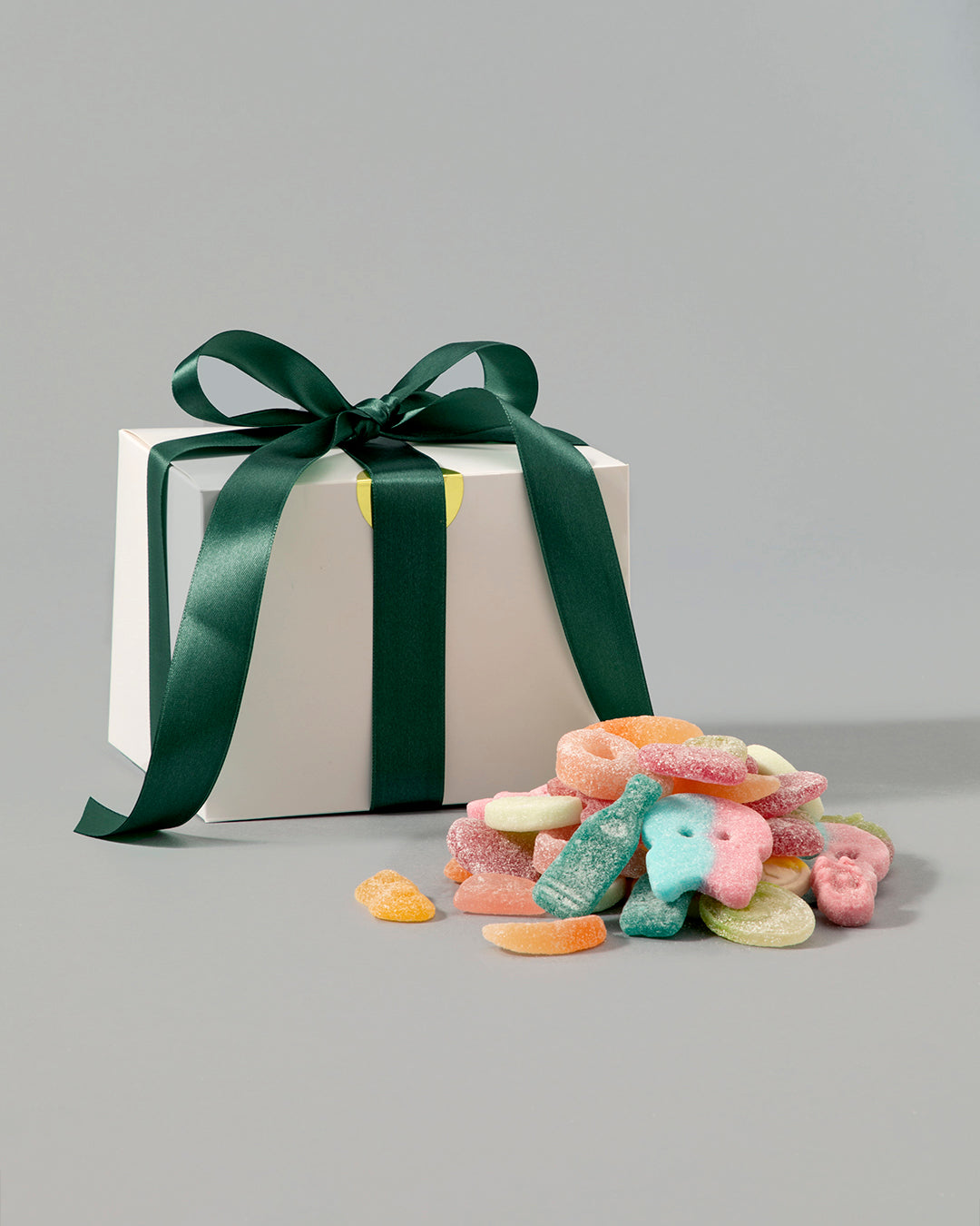 White gift box with dark green ribbon next to colorful vegan gummy candy mix on a gray background