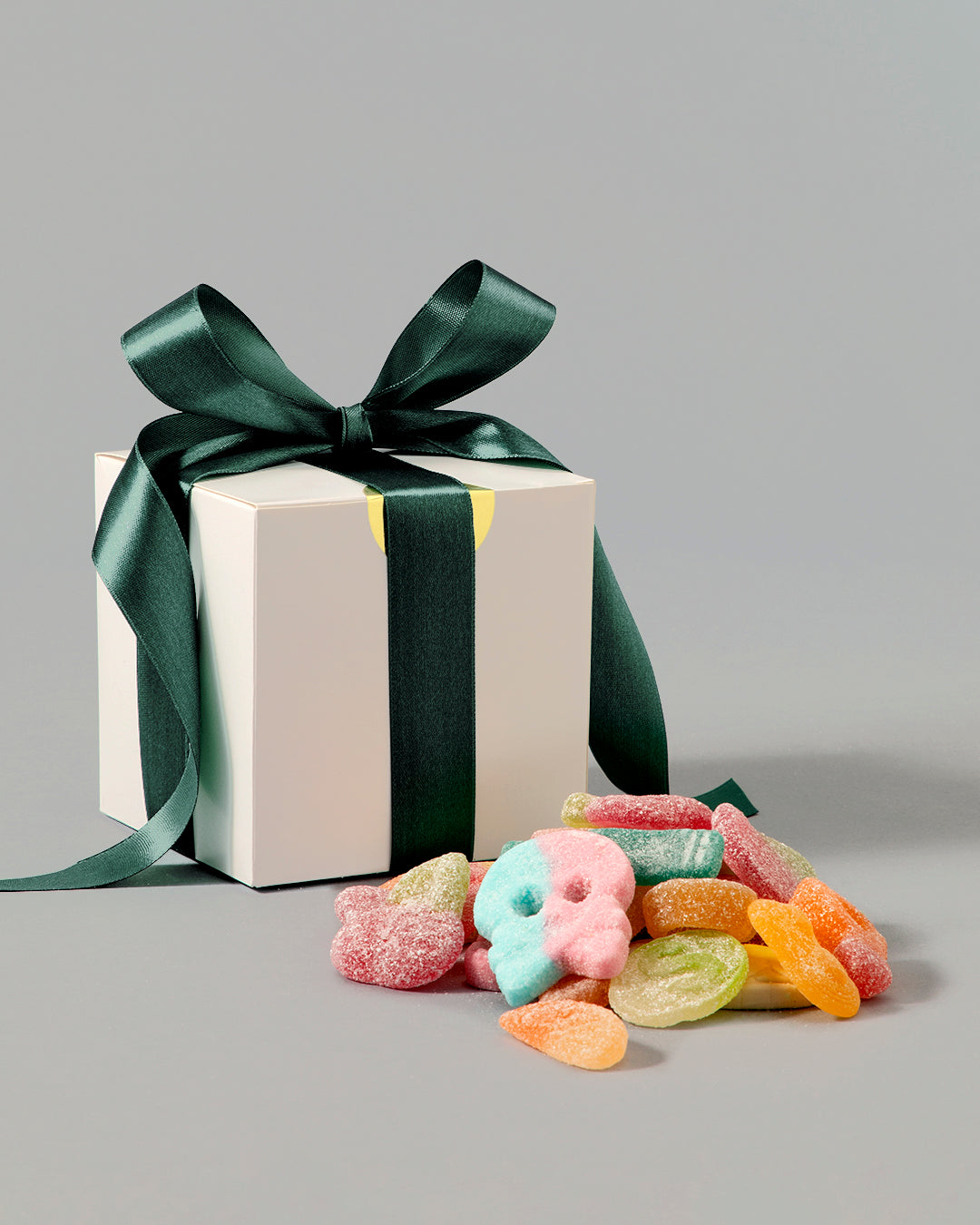 White gift box with dark green satin ribbon beside a colorful assortment of sugar-coated gummy candies on gray background