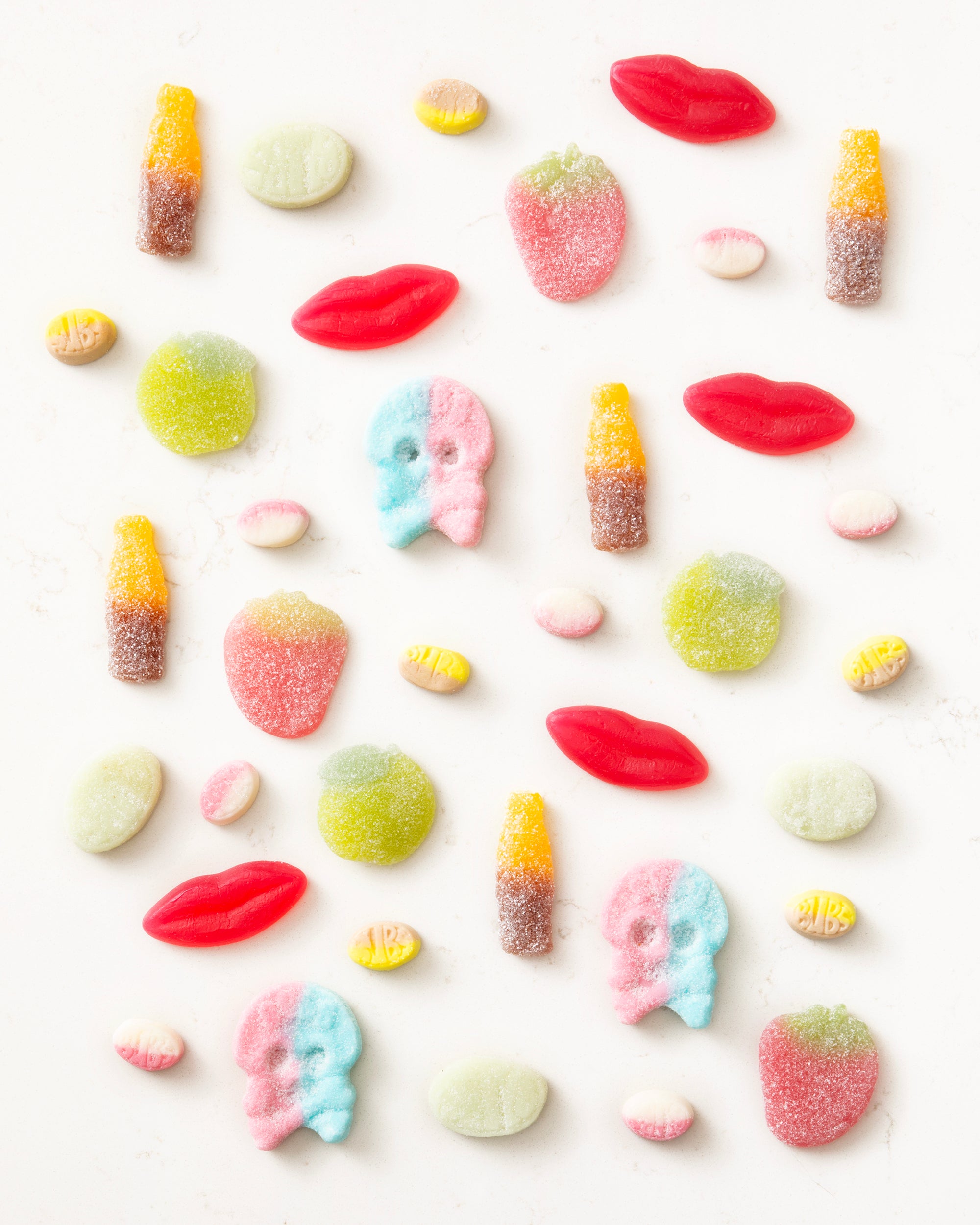 Assorted colorful vegan gummy candies shaped like lips, skulls, strawberries, bottles, and round discs on a white background